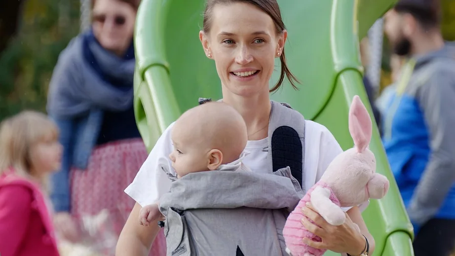 Une maman souriante dans un parc, son bébé porté face à elle dans un porte-bébé