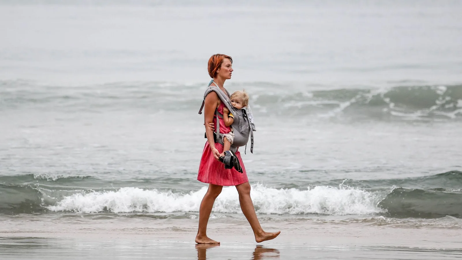 Une maman en robe marche au bord de la mer, son bébé porté face à elle dans un porte-bébé
